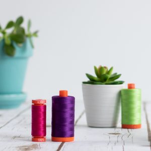 Style shot of three spools of cotton thread in purple green and red on white floor boards with a pot plant in the background