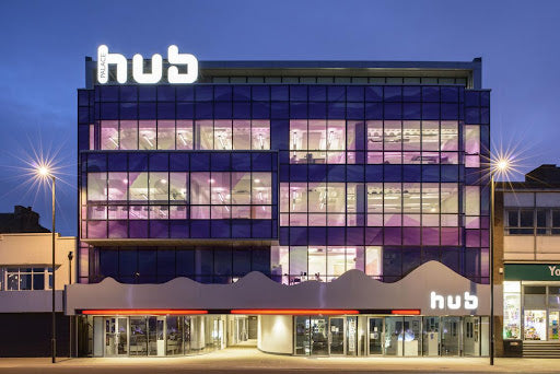 A photo of The Palace Hub, Redcar. A four storey building at dusk. The office windows are lit up, there are street lights at either side. There is a lilac tint in a wave shape on the bottom half of each window.